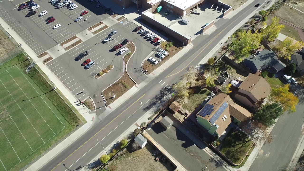 Overhead Hidden Lake, Colorado view with the parking lot and light-roofed building more centered, bordered by a curved road, houses, trees, and part of a green field, with no clearly visible people.