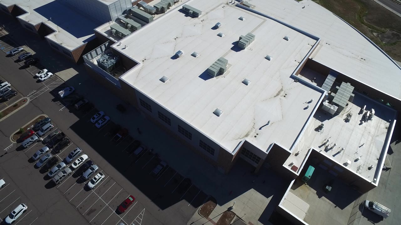 High aerial frame over a large white-roofed industrial building in Hidden Lake, Colorado, with rooftop units, parked cars along the left edge, and no visible people.