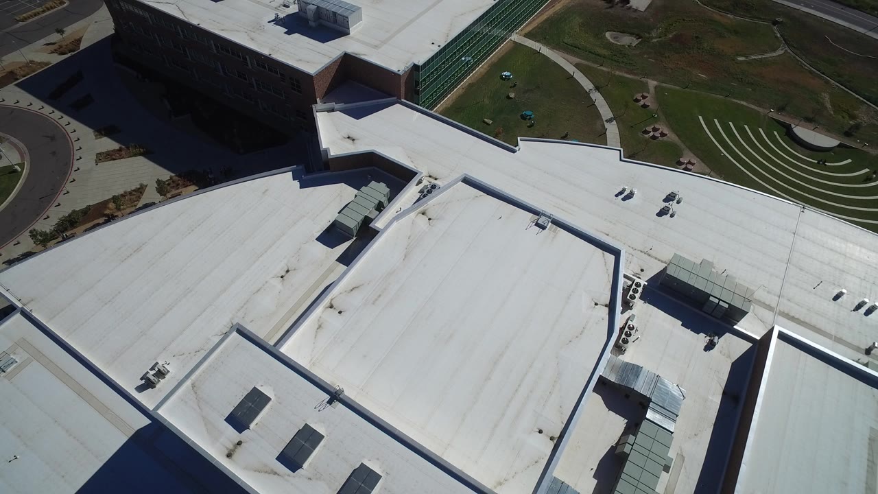 A steep aerial view in Hidden Lake, Colorado shows broad white rooftops, a dark building edge, a curved drive, and green lawn at the start of the clip, with no visible people.
