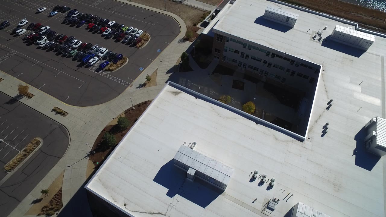 High oblique aerial frame in Hidden Lake, Colorado showing a white-roofed commercial building, rows of parked cars, sidewalks, and no visible people.