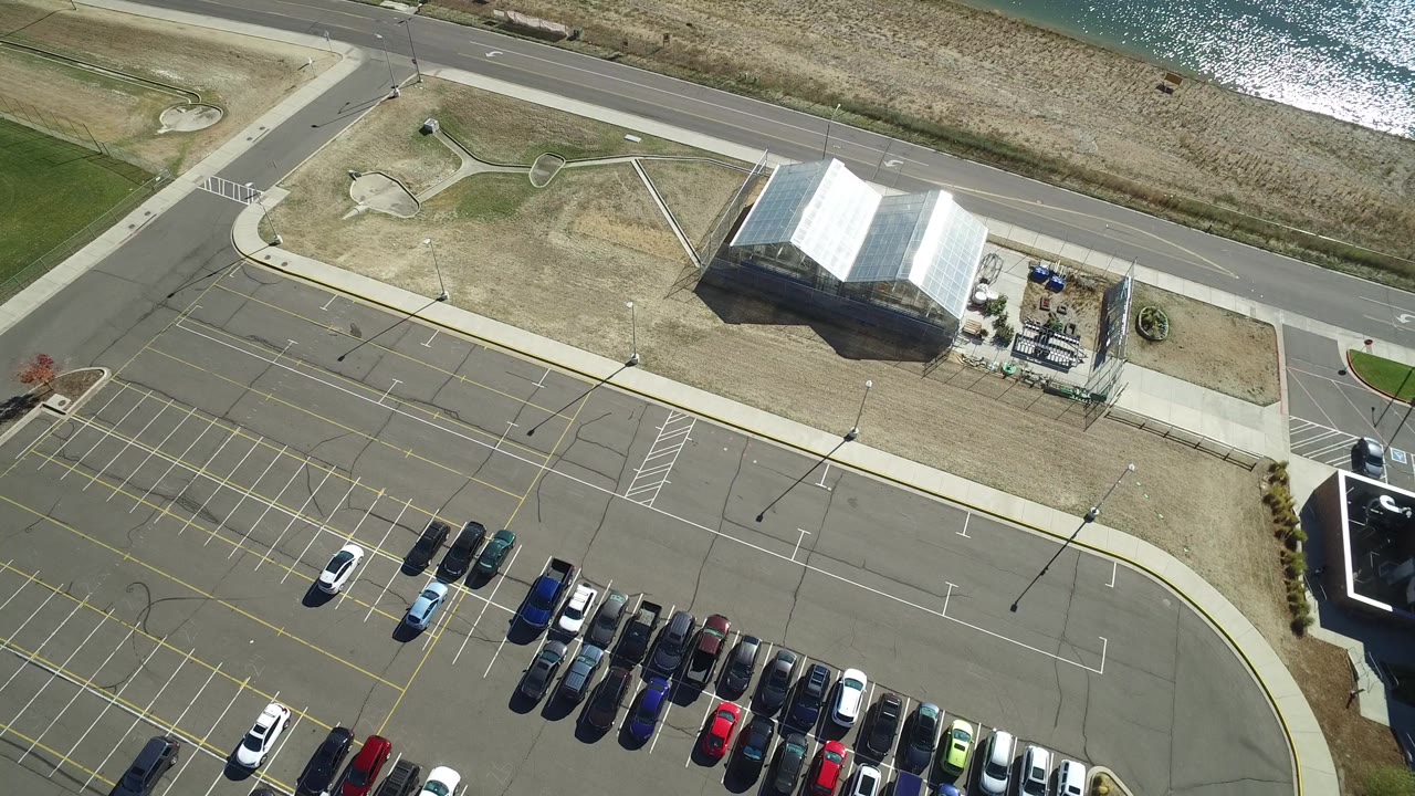 Aerial frame over Hidden Lake, Colorado with parked cars gathered along the lower parking rows, the road crossing the upper frame, and sunlight reflecting from the lake beyond the grass.