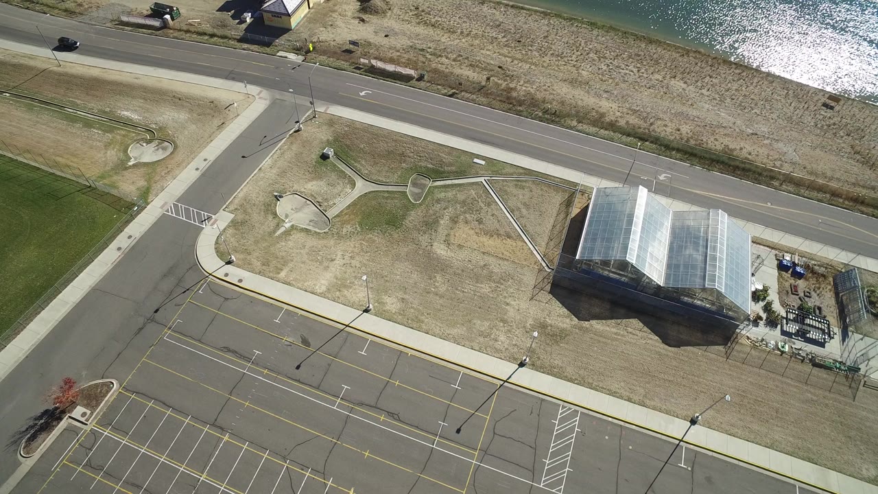 Aerial view of road, parking lot, dry field, glass-roofed building, and water.