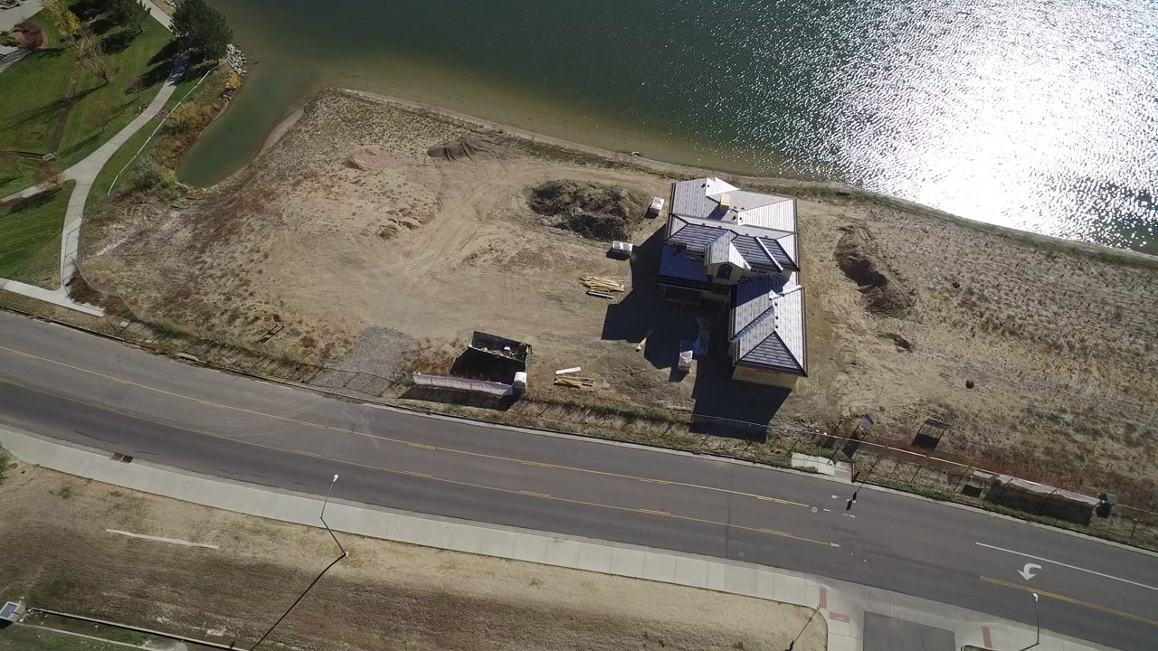 Overhead aerial frame in Hidden Lake, Colorado showing a sandy lakeside lot, a road and sidewalk at the bottom, a modern home near the shoreline, bright lake glare, and no visible people.