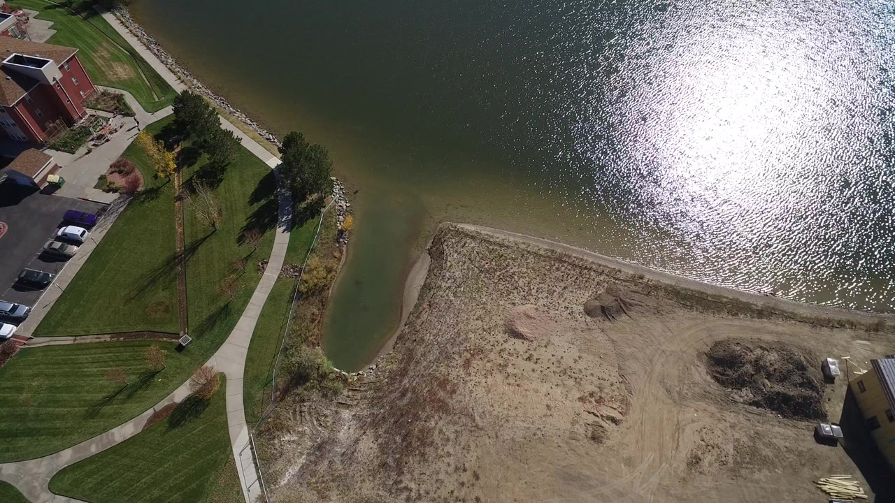 High aerial view over Hidden Lake, Colorado, showing sunlit green water, a curved shore, lawns, sidewalks, homes, parked cars, and bare dirt with no visible people.