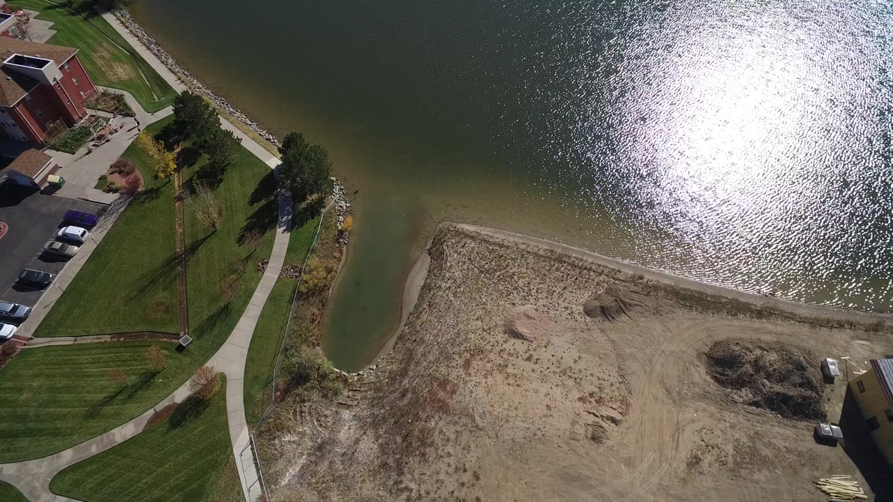 Aerial frame over Hidden Lake, Colorado, with a large bright reflection on the water, green lawns, a curved walkway, parked cars, homes, and no visible people.