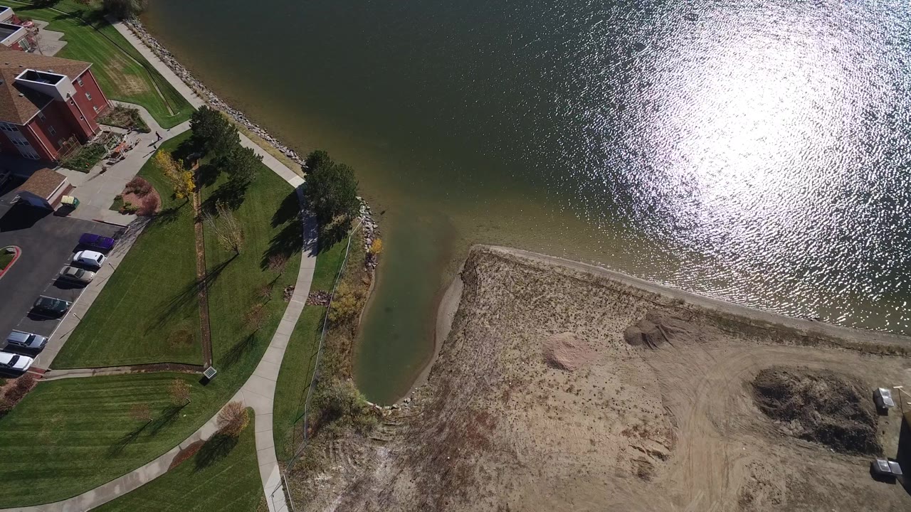 Aerial view of lake glare, residential building, parking area, lawns, sidewalk, and sandy dirt.