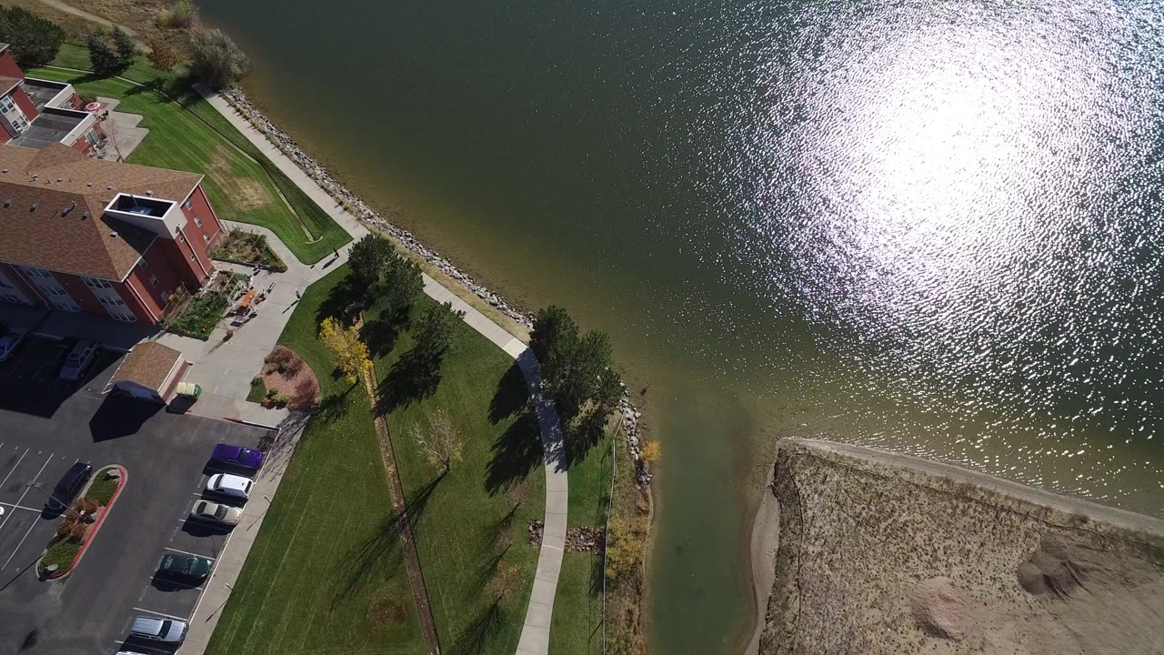 High aerial view of a lakeside building, lawn, walkway, parked cars, and sunlit lake water.