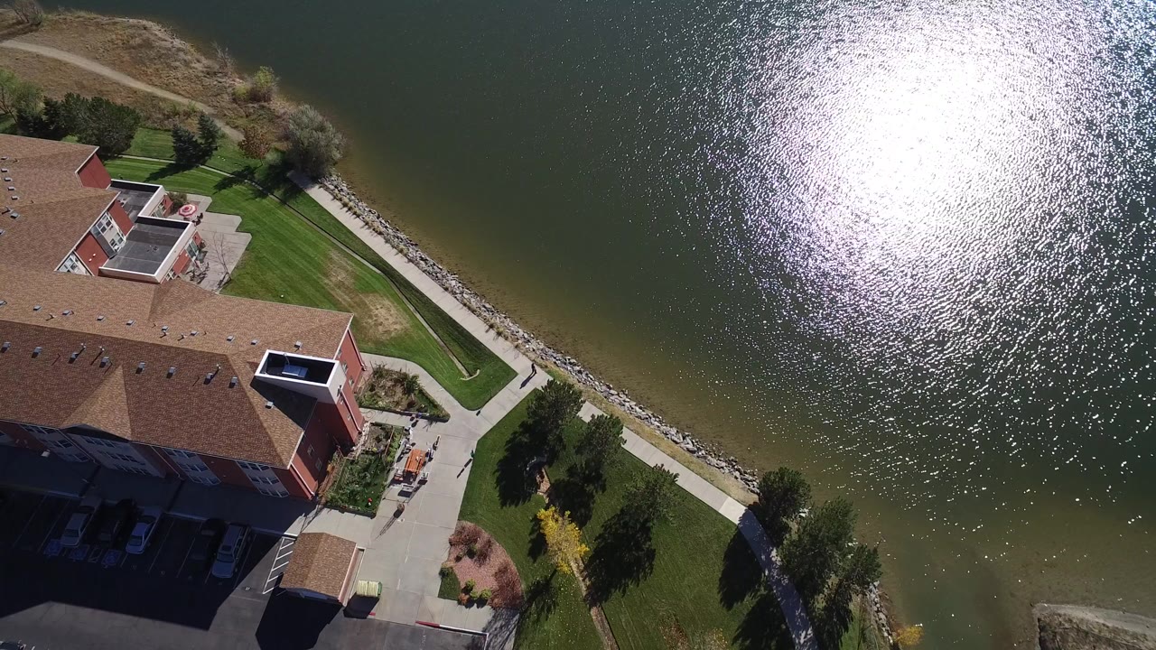 High aerial frame over Hidden Lake, Colorado showing a large brown-roofed building at lower left, green lawn, a rock shoreline, bright lake glare, and no visible people.