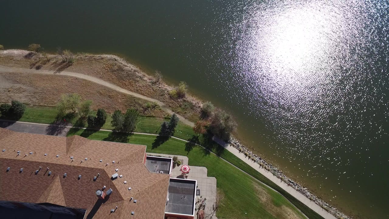 Overhead aerial frame in Hidden Lake, Colorado showing bright sun glare on dark lake water, a curved shoreline path, green lawn, trees, and the edge of a large house roof with no visible people.