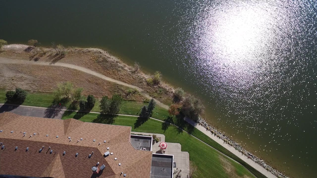 Top-down lakefront property view with roofline, trees, path, and shimmering water.