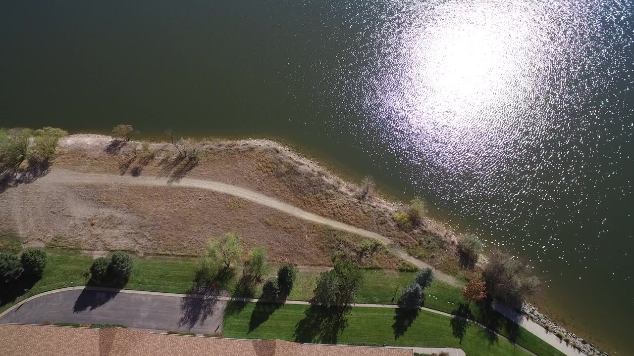 Aerial view over Hidden Lake, Colorado showing bright sun glare on dark water, a curving dirt shoreline path, scattered trees, grass, and no visible people.