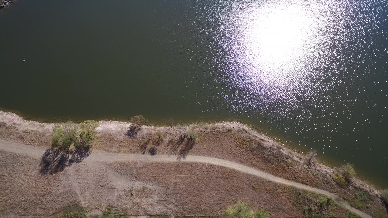 Overhead view of Hidden Lake, Colorado, with bright sun reflection on dark water, a pale dirt shoreline road, dry bank, scattered trees, and no visible people.