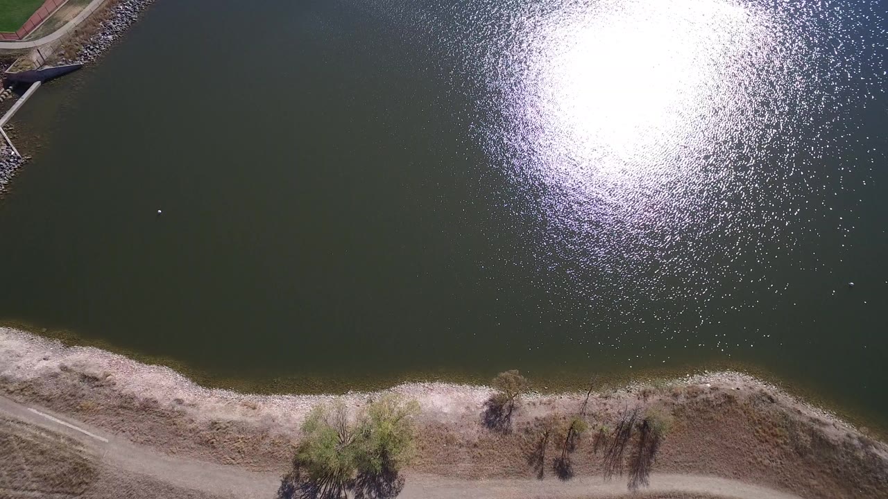 Overhead view of Hidden Lake in Colorado with dark green water, bright sun glare near the upper right, a pale sandy shoreline at the bottom, and no visible people.