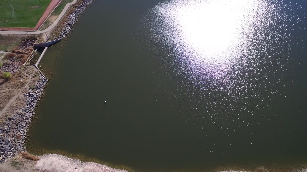 Aerial view over Hidden Lake, Colorado pond water with a large sun reflection near the upper right, rocky shoreline and paths at left, and no visible people.