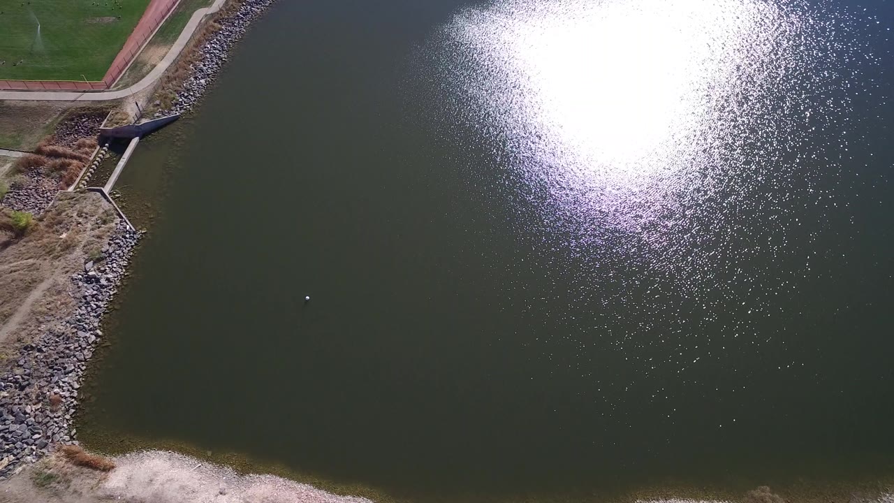 Aerial frame over Hidden Lake, Colorado showing dark pond water, bright glare on the upper right, grass and a curved path at the upper left, and no visible people.