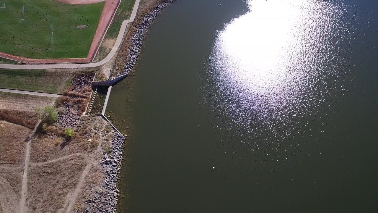 Overhead frame in Hidden Lake, Colorado showing a green sports field, shoreline rocks, a small water-edge structure, dark lake surface, bright sun reflection, and no visible people.