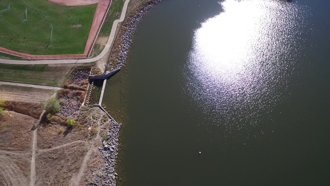 Top-down lake edge with a sports field, paths, rocky bank, and sun glare on water.