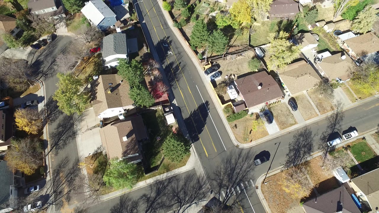 High overhead frame of Hidden Lake, Colorado homes beside a two-lane road, with parked vehicles, driveways, fenced yards, and long tree shadows.
