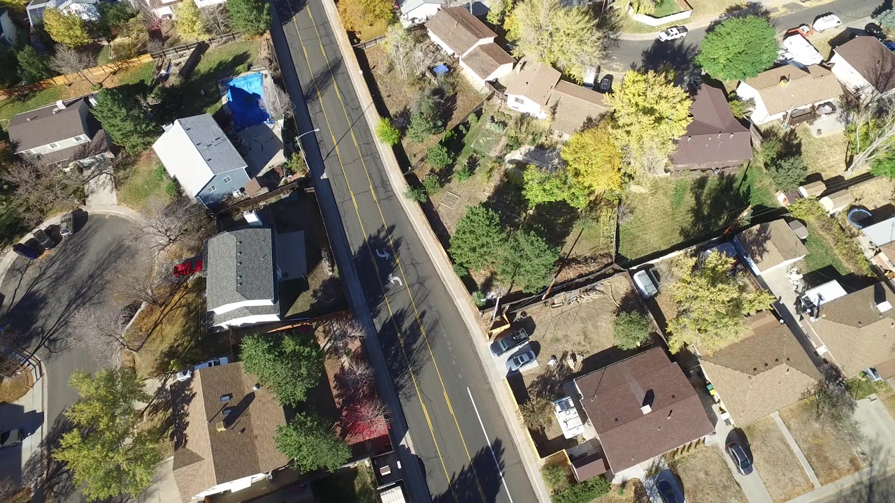 Top-down drone view of houses, yards, trees, parked cars, and a two-lane road.
