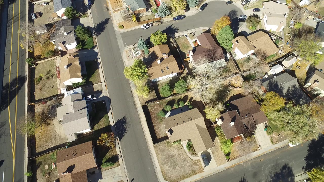 Overhead view of suburban homes, streets, yards, trees, and parked cars.