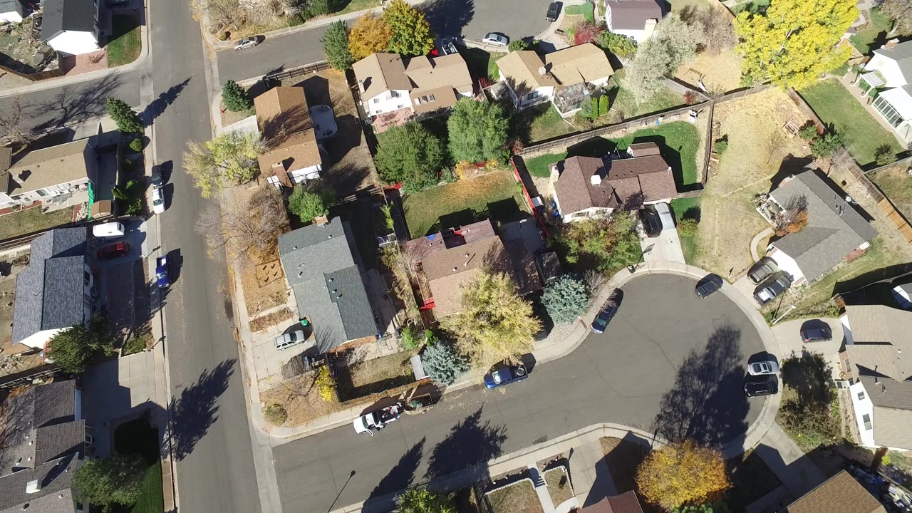 Aerial frame over Hidden Lake, Colorado showing a curved residential cul-de-sac, detached houses, driveways, yellow and bare trees, parked vehicles, and no visible people.