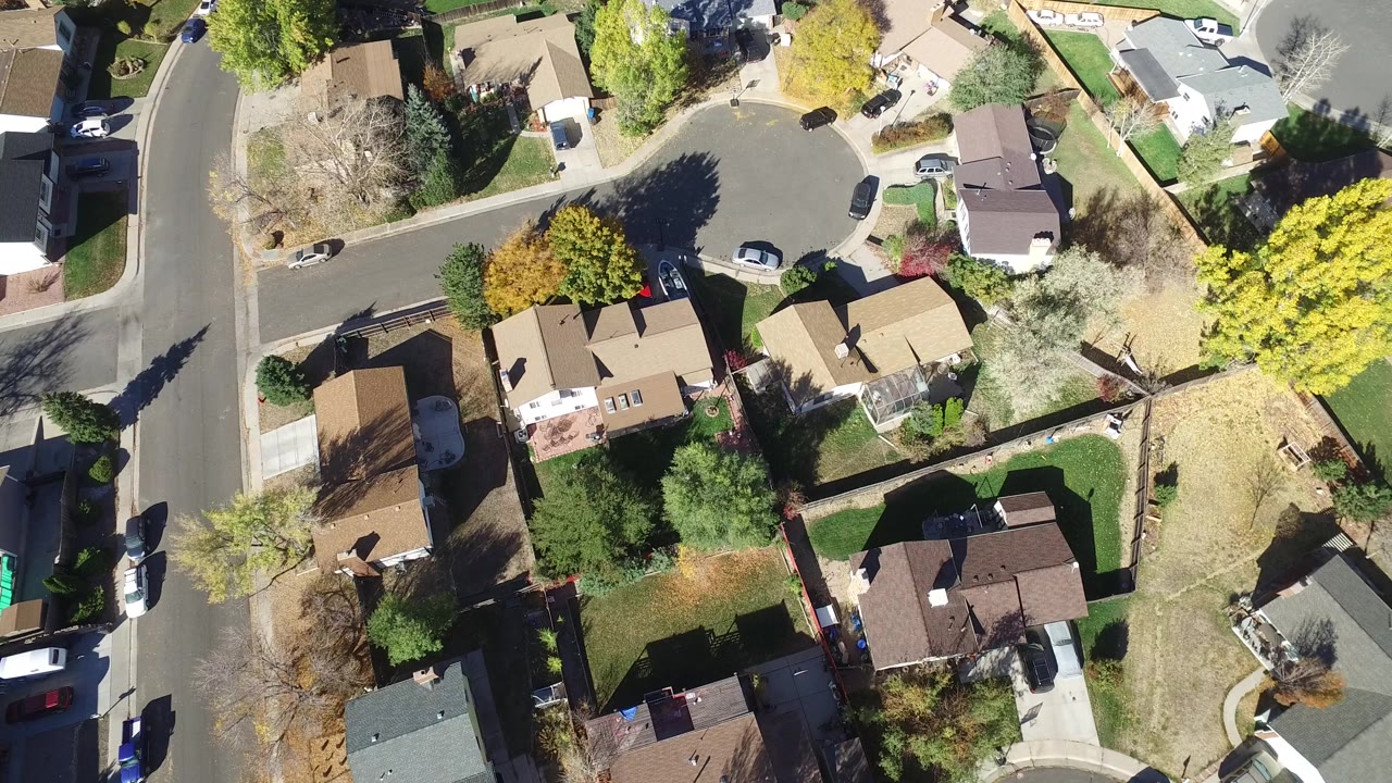 Overhead view of residential homes, curved streets, trees, lawns, and parked cars.