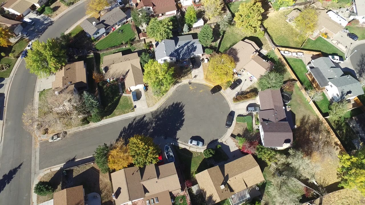 Bird's-eye frame over Hidden Lake, Colorado showing a broad street bend, clustered rooftops, green yards, autumn trees, parked cars, and no visible people.