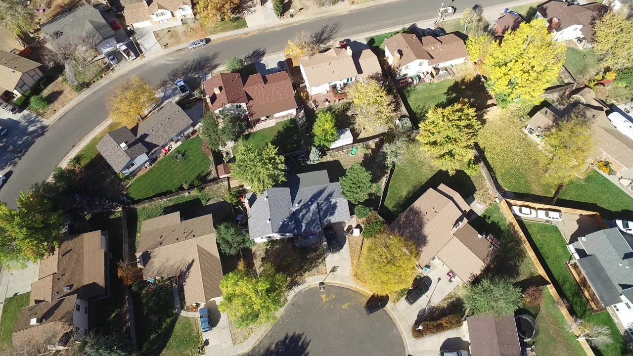 Overhead Hidden Lake, Colorado frame with the curving street near the top, clustered rooftops below, green lawns, fenced backyards, and sunlit yellow trees.