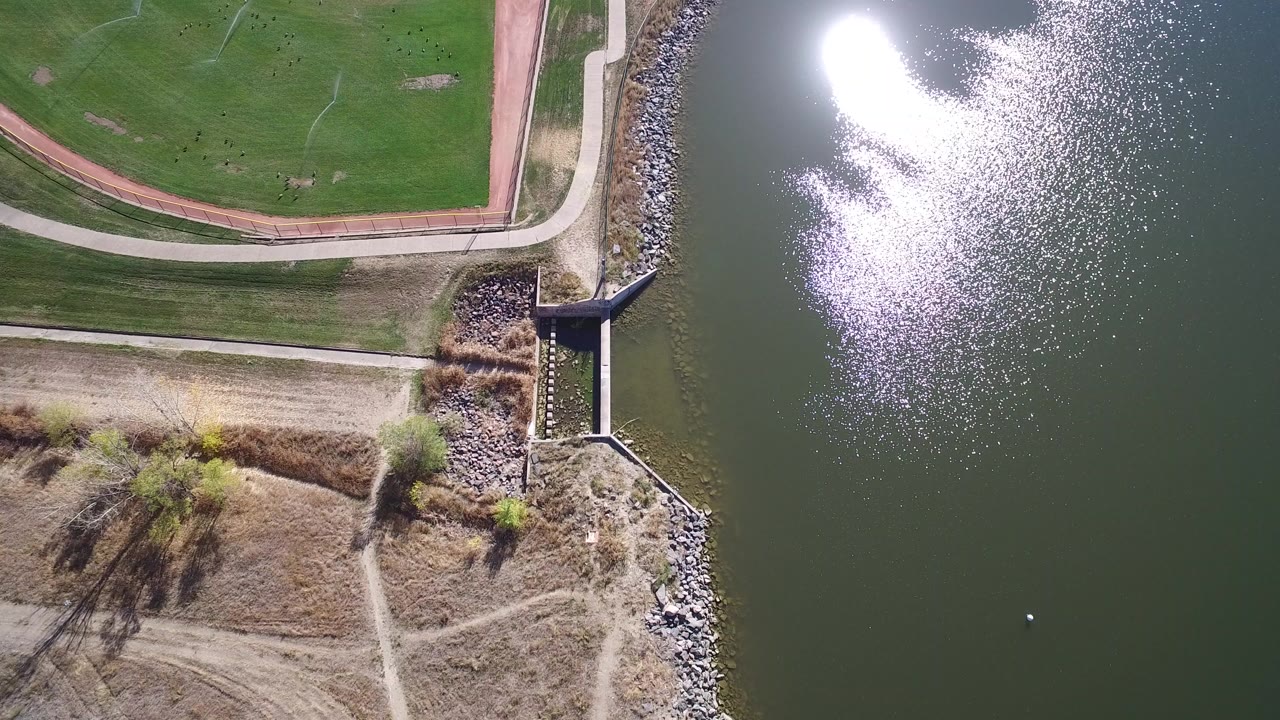 Overhead drone frame at Hidden Lake Colorado showing the concrete spillway between riprap shoreline and green water, with dirt paths, grass, bright water reflection, and no visible people.