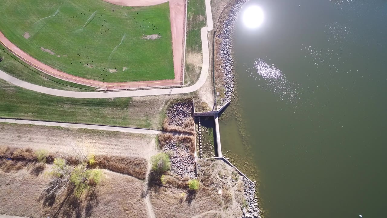 Top-down aerial frame over Hidden Lake, Colorado showing the corner of a baseball field, a pale path, riprap shoreline, a drainage outlet, dark lake water with sun glare, and no visible people.