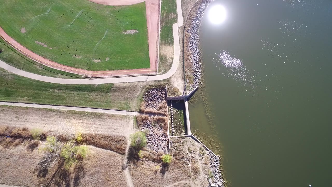 Aerial frame over Hidden Lake, Colorado with the baseball field shifted slightly left of center, the curved path above the drainage outlet, shoreline rocks, lake water, bright glare, and no visible people.