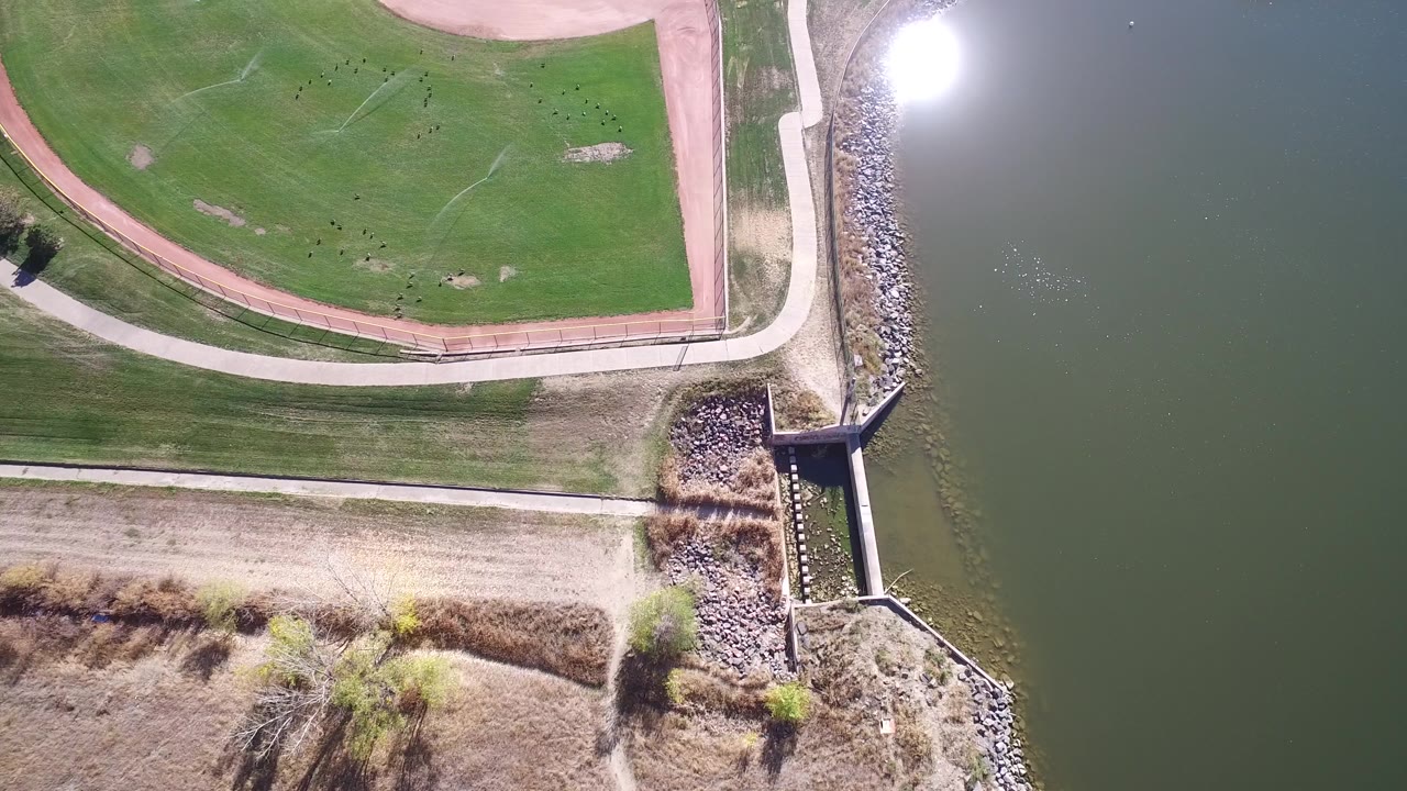 Overhead frame from Hidden Lake, Colorado showing golf course grass, a pale path, rock revetment, and green water with bright sun reflection, no visible people.
