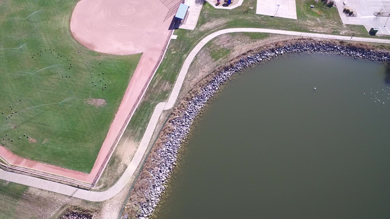Overhead frame at Hidden Lake Colorado showing dark green water, a curved rock shoreline, park paths, grass, a pink ballfield, and no visible people.