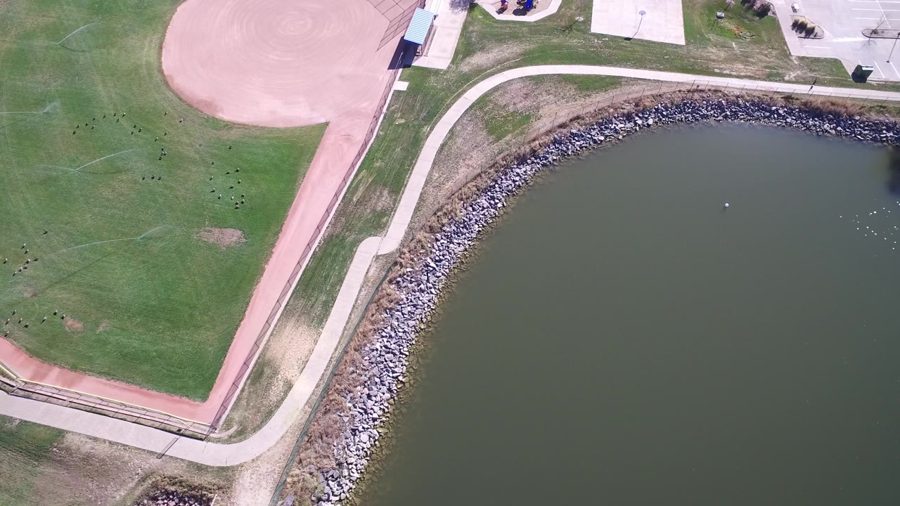 Aerial frame at Hidden Lake Colorado with the curved path more centered between grass, the rock border, lake water, ballfield dirt, and no visible people.