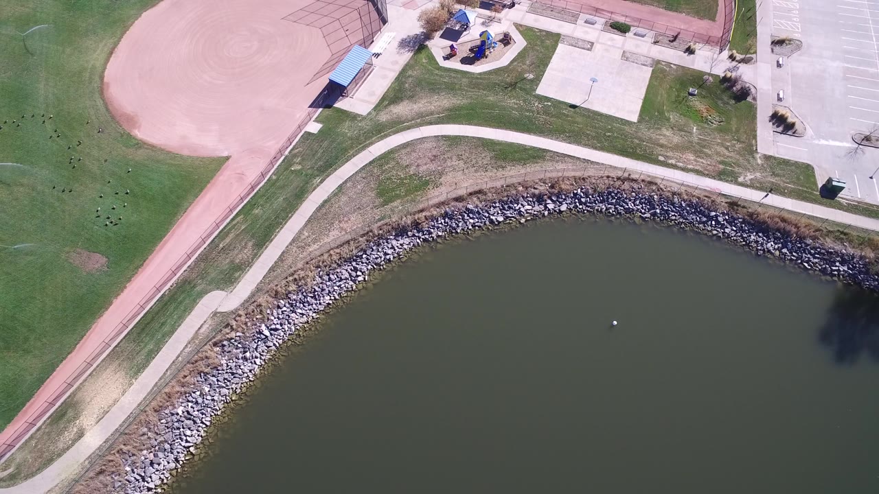 High aerial frame over Hidden Lake, Colorado showing green pond water, curved rock shore, walking path, baseball infield, playground area, and no visible people.