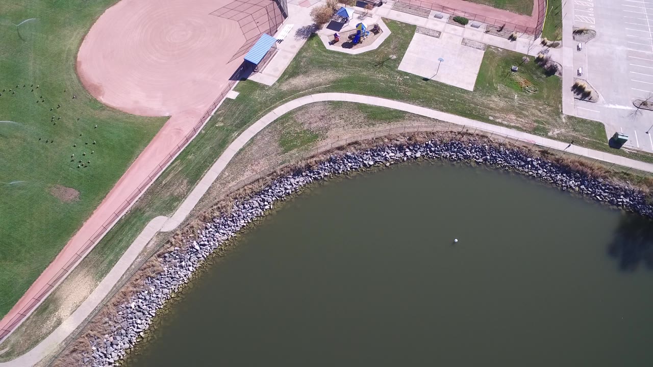 Overhead view in Hidden Lake, Colorado with the pond across the lower half, a stone-lined bank, pale path, ball diamond corner, play area, parking spaces, and no visible people.