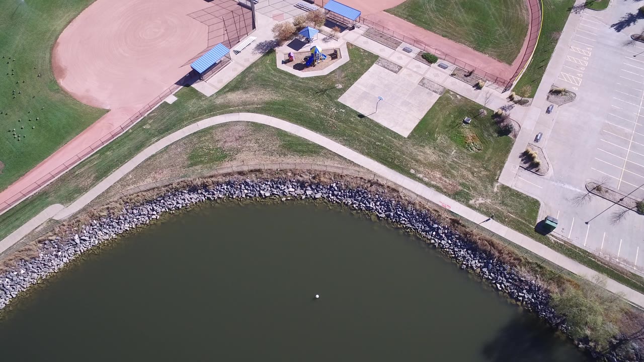 Top-down aerial frame over Hidden Lake, Colorado, showing dark lake water, a curved rock shoreline, pale walking path, baseball field edge, playground area, blue shelters, an empty parking lot, and no visible people.