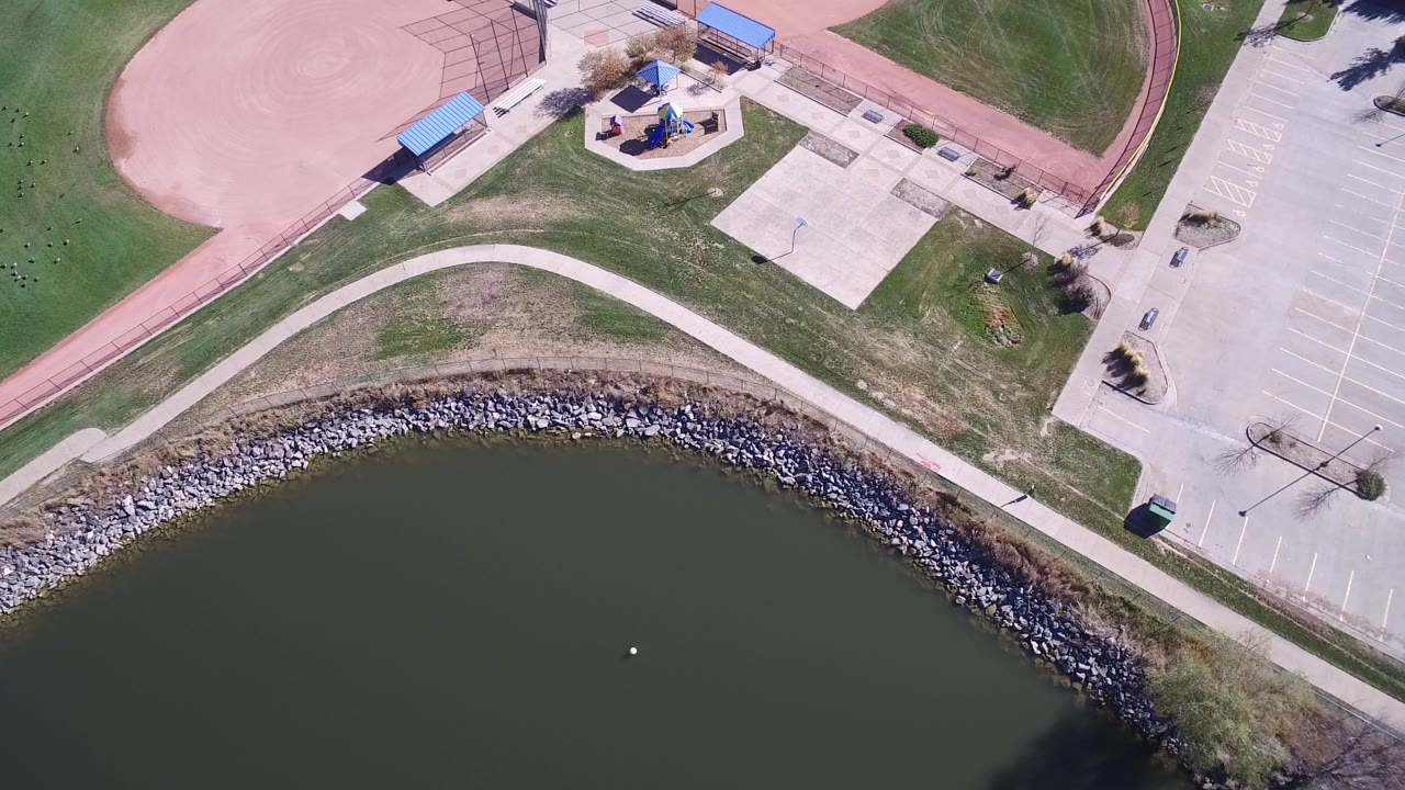 Overhead park view with lake water, riprap shoreline, path, playground, and parking area.