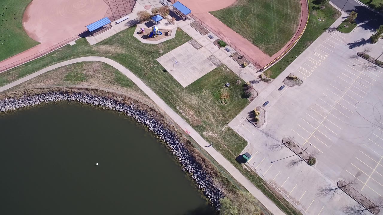 Aerial view of lake edge, sidewalk, parking lot, and baseball field corner.