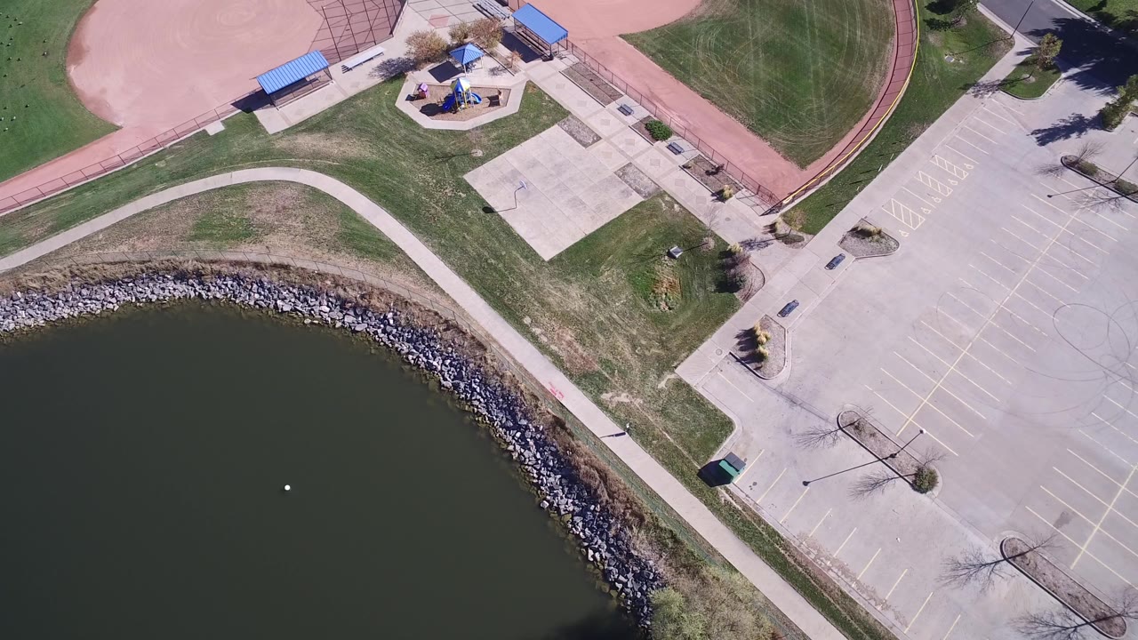 Aerial frame in Hidden Lake, Colorado with the lake occupying the lower left, riprap shoreline, a broad empty parking lot, grass medians, and no visible people.