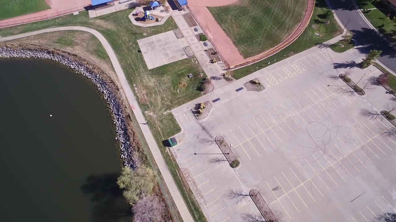 Opening overhead frame in Hidden Lake, Colorado showing dark lake water, a rock shoreline, an empty parking lot, paths, grass, and no visible people.