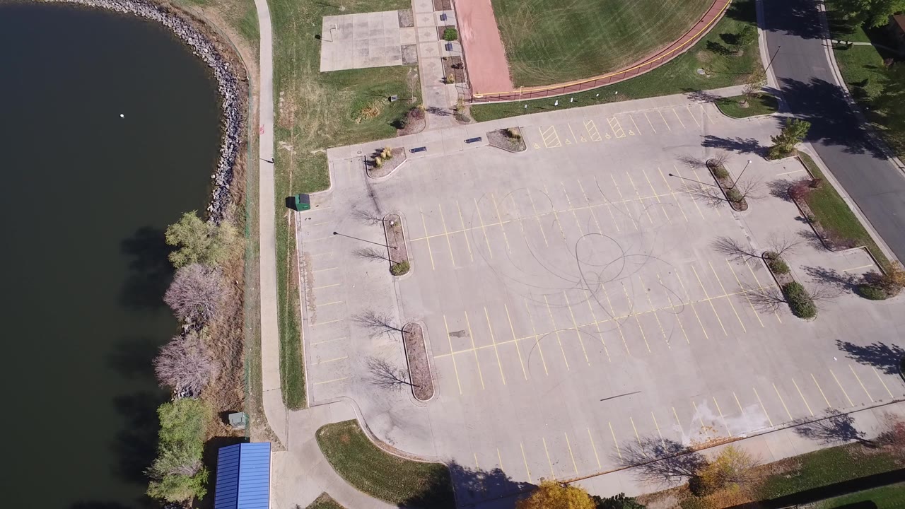 Overhead aerial frame in Hidden Lake, Colorado showing a mostly empty marked parking lot beside dark lake water, with grass, trees, paths, and no visible people.