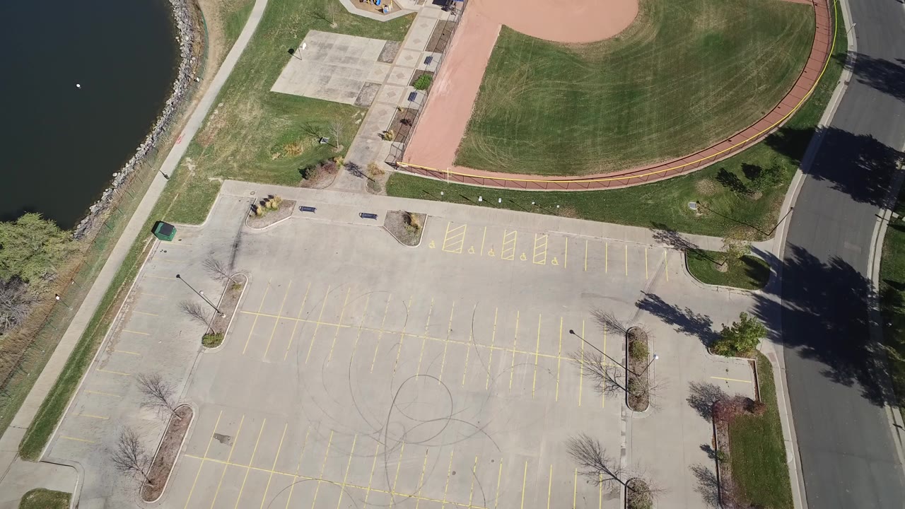 High aerial frame over Hidden Lake, Colorado showing part of a baseball field, an empty striped parking lot, a road, lakeside path, and no visible people.