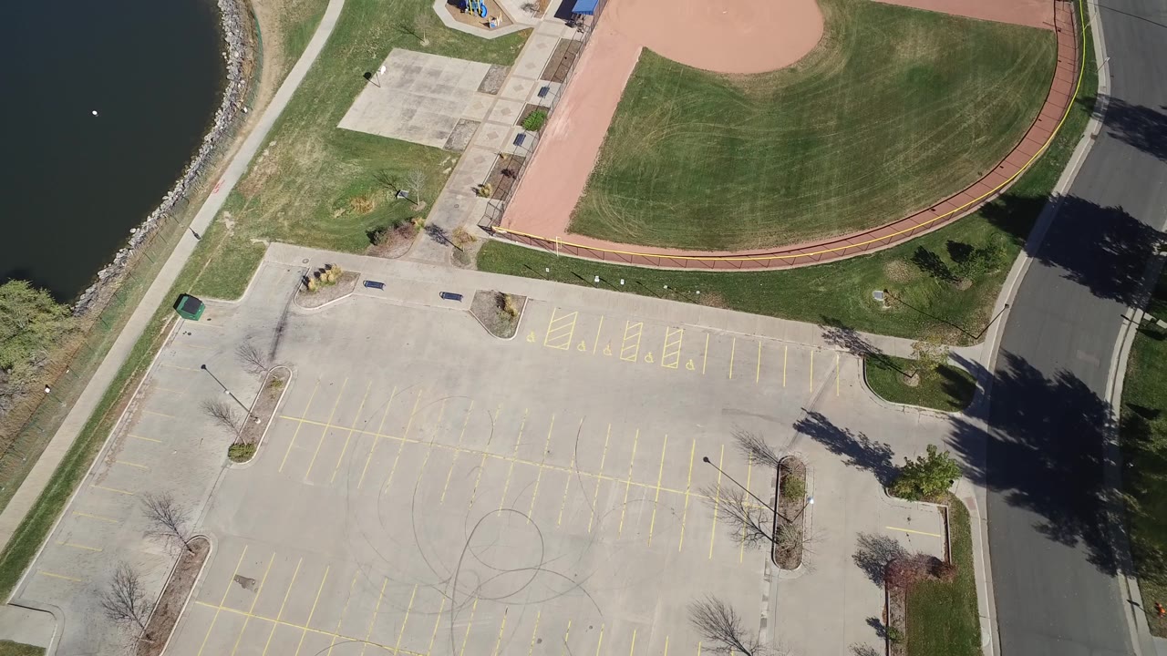Aerial frame of a sports diamond, parking lot, paths, and roadside shadows.
