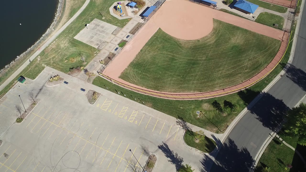 Overhead aerial frame at Hidden Lake, Colorado showing the curved green ballfield, tan infield dirt, paved court area, parking lines, road edge, shoreline water, and no visible people.