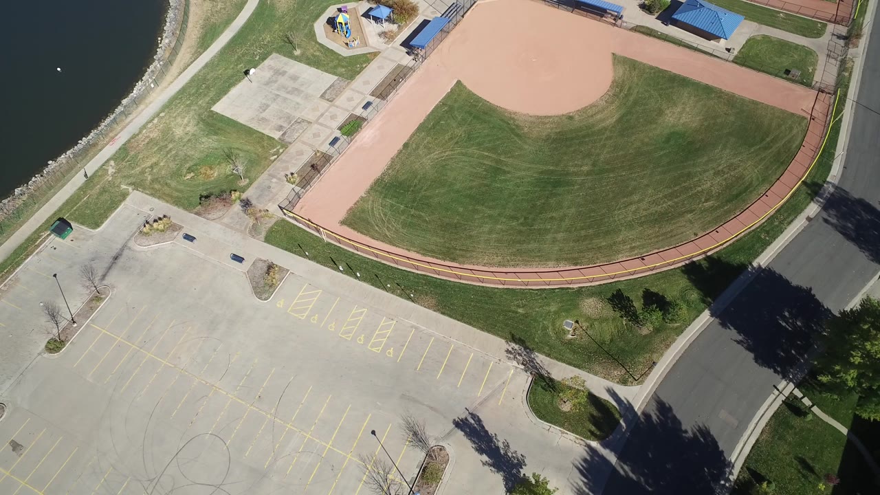 Aerial frame at Hidden Lake, Colorado with the ballfield centered more tightly, the shoreline along the left edge, paved recreation areas below, blue-roofed structures near the top, and no visible people.