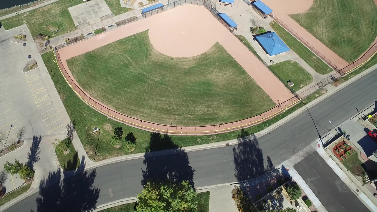 Overhead frame from Hidden Lake, Colorado showing a curved residential road below a ballfield, green lawns, paved paths, parking lots, blue-roof structures, tree shadows, and no visible people.