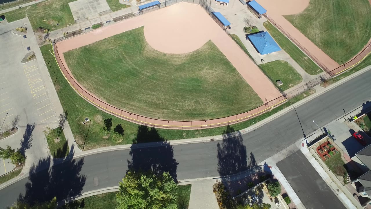Aerial view of a residential road below a baseball field and green open space.