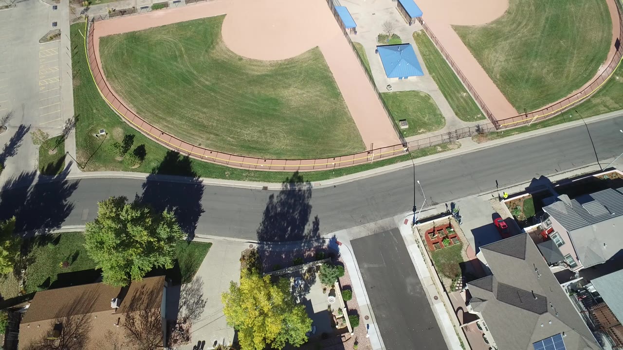 Aerial frame in Hidden Lake, Colorado with the park path arc high in frame, a two-lane street across the center, and residential roofs and yards along the lower edge, no visible people.