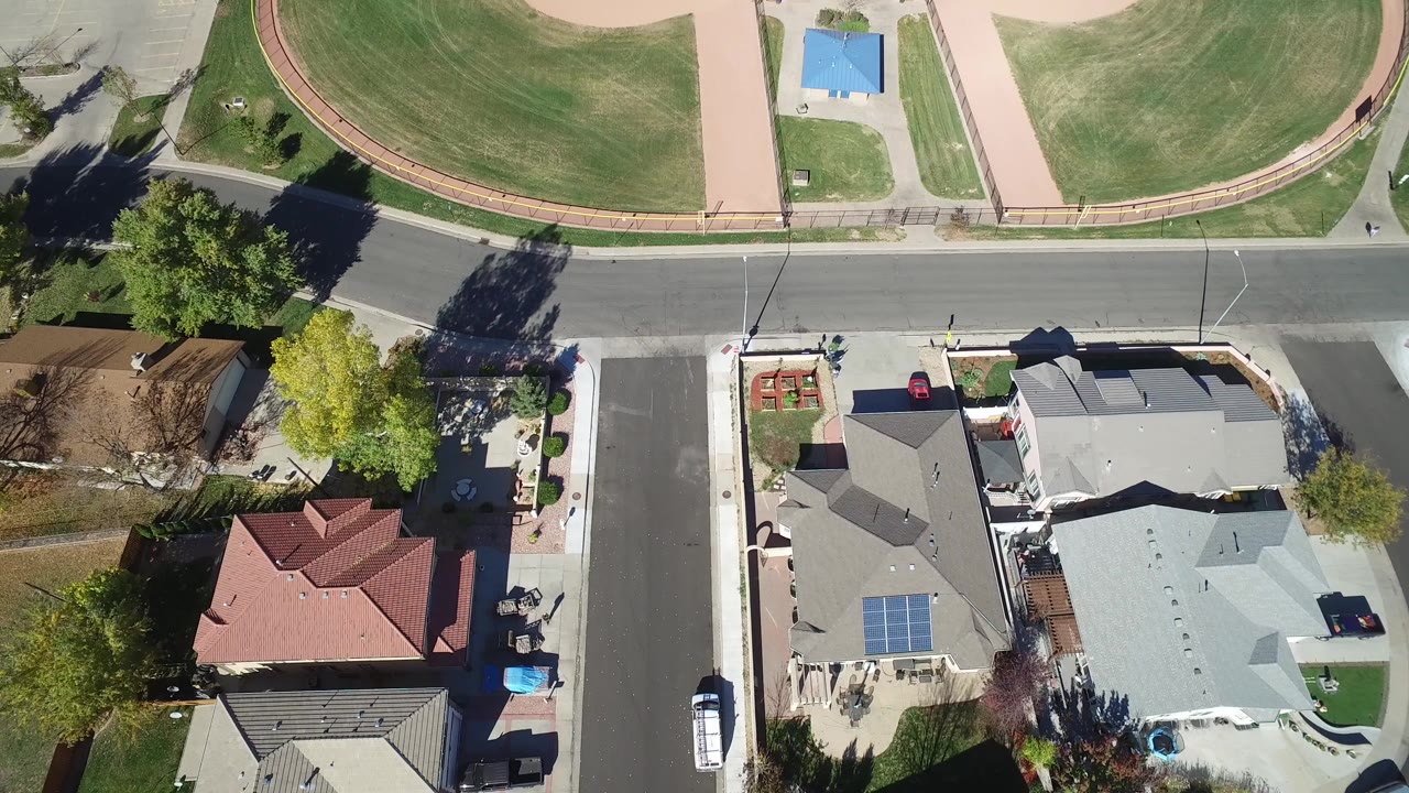 Opening overhead frame in Hidden Lake, Colorado showing detached homes, a paved street, lawns, trees, and a green recreation area across the road with no visible people.